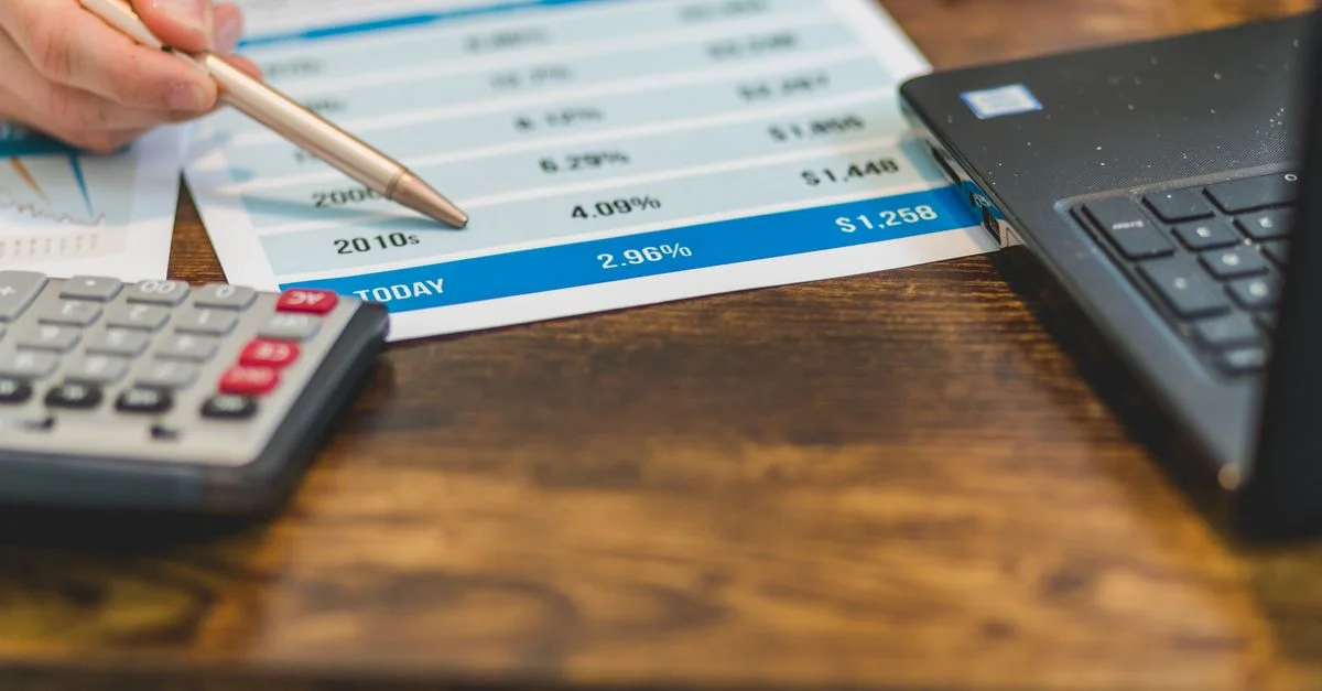 Gros plan d'une main avec un stylo analysant des taux financiers sur du papier avec une calculatrice et un ordinateur portable à proximité.